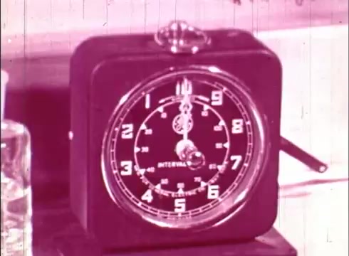 The still features a vintage black clock with a prominent face, marked with bold numbers and an adjustable hand. The clock appears to be a timer, indicated by the word 'INTERVAL' on its display. It is set against a simple background, likely in a laboratory or educational setting, alongside a container, hinting at a scientific context related to cellular studies. The image is tinted in a pinkish hue, typical of older film prints.