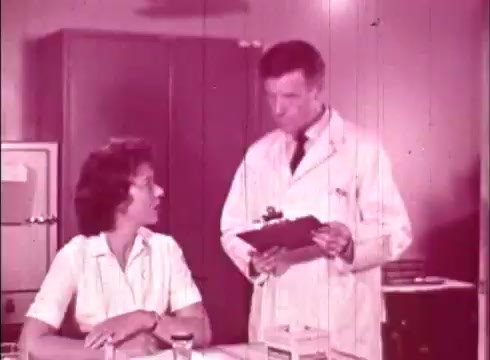 The still depicts a scene from the educational film 'The Human Cell and the Cytologist.' It features a male cytologist in a lab coat, standing and holding a clipboard, while a female subject sits at a table, attentively looking at him. The setting appears to be a laboratory, indicated by the visible equipment and cabinets in the background. The overall tone is informative, likely focusing on a discussion related to cellular biology. The image shows the characteristic graininess and color palette typical of 16mm film.