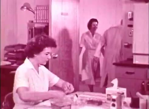 The still from 'The Human Cell and the Cytologist' features a laboratory setting with a woman in a white lab coat seated at a table, engaged in scientific work. She appears to be handling materials, possibly preparing samples. In the background, another woman dressed in a light-colored dress enters the frame, while a third figure in a lab coat is partially visible. The overall tone of the image is pinkish, indicative of the film's vintage aesthetic. The scene exemplifies an educational environment focused on cytology.