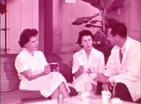 The still from 'The Human Cell and the Cytologist' depicts three individuals engaged in conversation. Two women in white attire sit together, one holding a cup, while a man in a lab coat sits across from them. The background suggests a relaxed environment with greenery, likely part of a casual educational setting. The color tone is predominantly pinkish, typical of certain 16mm films. Their conversation likely pertains to cytology and human cells, contributing to the film's educational theme.