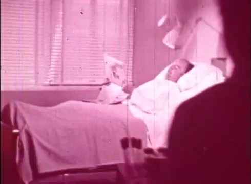 The still from 'The Human Cell and the Cytologist' shows a patient lying in a hospital bed, reading a newspaper. The scene is bathed in a reddish hue typical of older 16mm prints. Sunlight filters through blinds, creating a serene atmosphere, while the presence of medical equipment suggests a focus on health and treatment. This educational film likely illustrates the relationship between human cells and medical environments.