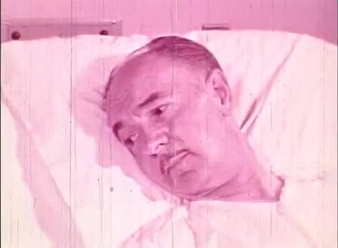 The still features a middle-aged man lying in a hospital bed, appearing contemplative. The scene is marked by a pink tint typical of older 16mm films, suggesting a warm, educational tone. The context indicates it is part of an informational film focused on human biology, likely discussing cellular processes or health-related themes. The man's expression may convey a sense of inquiry or reflection relevant to the film's educational purpose.