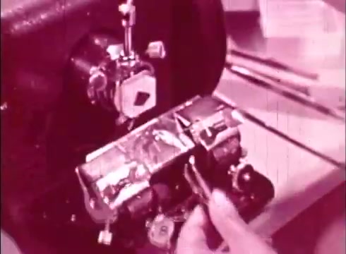 The still shows a close-up of a scientific device or apparatus, likely a cytological instrument, being handled by an individual. The scene is in a muted pink tone, characteristic of older 16mm prints, emphasizing a laboratory setting. The focus is on the precision and detail involved in studying cells, reflecting the educational purpose of the film.