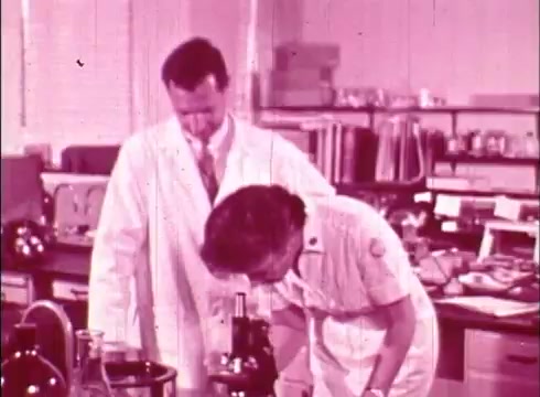 The still shows two figures in a laboratory setting, engaged in a scientific activity. One person, in a white lab coat, stands closely beside another, who is looking through a microscope. The environment is filled with various lab equipment and glassware, typical of a cytology or biological research setting. The overall tone is educational, likely focusing on cellular studies and cytology processes. The film is characterized by a vintage look due to the 16mm format.