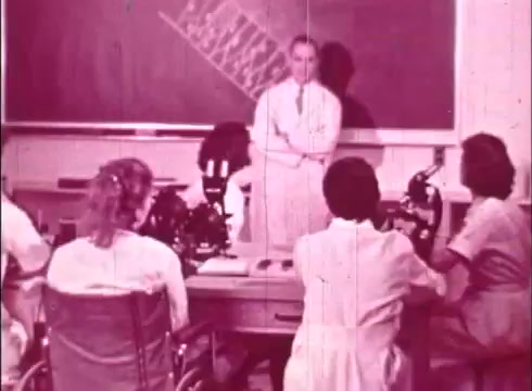 The still from the educational film 'The Human Cell and the Cytologist' features a classroom setting. A female instructor stands in front of a chalkboard, engaging with students. The students, seated at lab tables with microscopes, appear focused on the lesson. The image has a vintage quality, typical of 16mm prints, and emphasizes a scientific learning environment.
