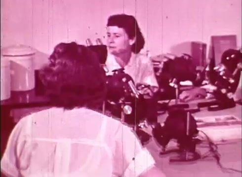 The still from 'The Human Cell and the Cytologist,' likely an educational film, features two women in a laboratory setting. One woman is seated at a desk with microscopes, appearing focused and engaged, while the other, partially visible in the foreground, has her back to the camera. The environment is well-lit, with a background filled with scientific equipment and books, emphasizing an academic atmosphere. The color tone suggests a vintage style characteristic of 16mm film.