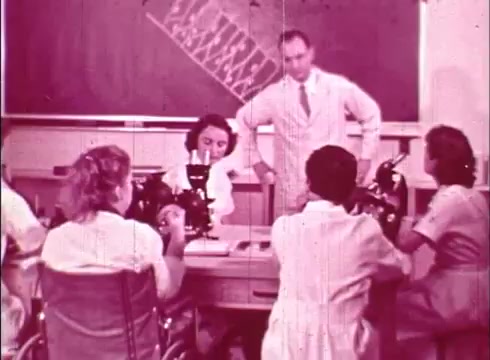 The still from 'The Human Cell and the Cytologist' depicts a classroom scene focused on cytology. Central to the composition is a male instructor standing, engaged in discussion with four students seated around a table, each using microscopes to examine specimens. The environment suggests an educational setting, with a chalkboard in the background, likely featuring diagrams or notes related to cellular study. The film's vintage quality is evident, reflecting the era of 16mm educational films.