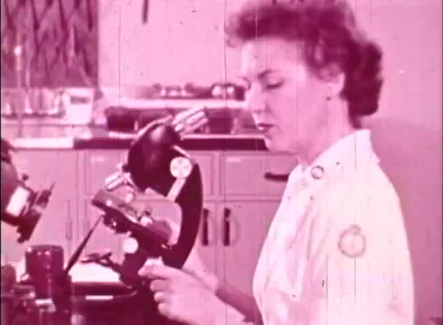 The still from 'The Human Cell and the Cytologist' features a woman in a lab coat, engaged in microscopy work. She appears focused as she examines a specimen under a microscope. The image has a vintage quality typical of 16mm prints, with a pinkish tint, suggesting it's part of an educational film aimed at discussing cellular biology and cytology. The lab environment includes various laboratory equipment visible in the background.
