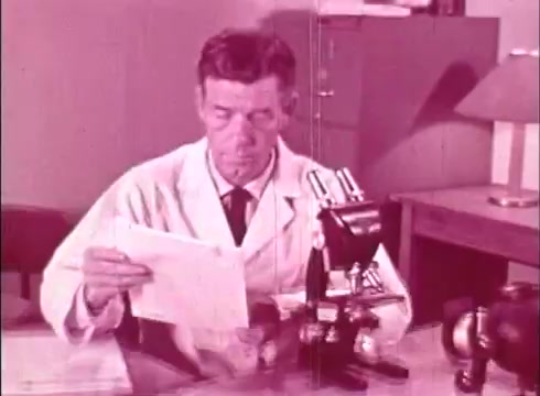 The still from 'The Human Cell and the Cytologist' depicts a man in a white lab coat sitting at a table, examining documents while looking thoughtfully at a piece of paper. In front of him is a microscope, suggesting a focus on scientific study, likely related to cytology. The background includes a lamp and filing cabinet, indicating a laboratory setting. The overall color tone is pinkish, characteristic of vintage educational films.