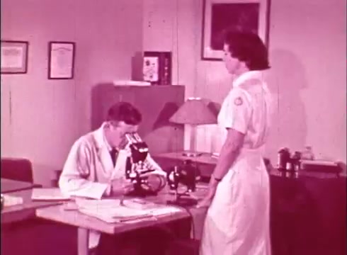 The still depicts a scene from an educational film titled 'The Human Cell and the Cytologist.' In a laboratory setting, a male scientist dressed in a white lab coat is focused on a microscope, likely examining a specimen. Standing nearby is a female nurse or assistant in a white uniform, attentively engaged in conversation with him. The environment is clinical, with certificates and scientific equipment visible in the background, creating a context conducive to educational discourse about cellular biology.