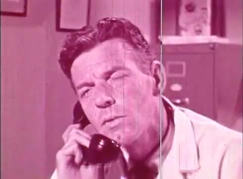 The still from 'The Human Cell and the Cytologist' depicts a man in a lab coat intently listening while on the phone. The image has a pinkish hue typical of 16mm film prints, and the background suggests a laboratory setting with filing cabinets. The expression on his face indicates concentration, likely engaged in a discussion related to cytology or cellular biology.