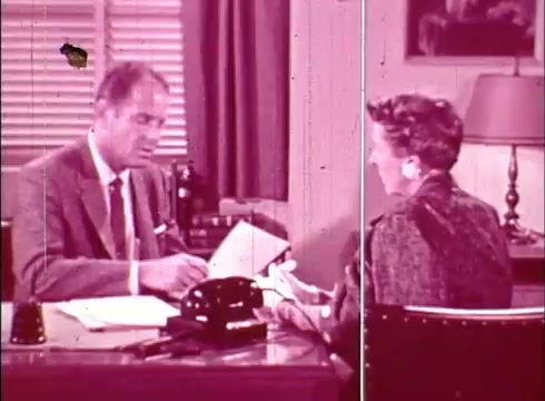 The still shows a mid-century office setting with two individuals engaged in a discussion. A man, dressed in a suit, is seated at a desk, examining documents, while a woman, wearing a stylish jacket, listens attentively. The room features a lamp, a typewriter, and partially closed blinds, indicating a professional environment typical of educational films from that era. The color tones are slightly faded, characteristic of 16mm film prints.