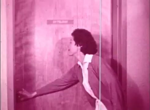 The still shows a woman approaching a door, likely in a laboratory or educational setting. She appears engaged, possibly knocking or about to enter. The image is presented in a pink hue, typical of the period's film stock, and suggests an instructional context related to cytology. The door is labeled, which adds to the educational theme of the film.