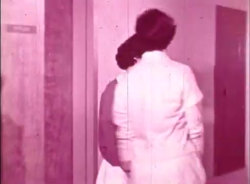 The still from 'The Human Cell and the Cytologist' depicts two individuals walking in a corridor, likely within a laboratory or educational setting. The image is presented in a pink hue, characteristic of older 16mm prints, suggesting a focus on the subjects rather than vibrant color. The ambiance hints at an instructional purpose, possibly leading to a demonstration or discussion about cytology. The presence of lab coats indicates a scientific context, emphasizing professionalism.