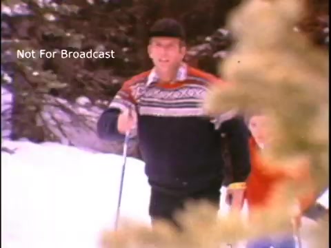 The still from the John Denver PSA features him skiing in a snowy landscape, wearing a colorful sweater and a hat. He appears cheerful and engaged in outdoor physical activity, promoting fitness and a healthy lifestyle. A child can be seen skiing alongside him, reinforcing the message of encouraging physical activity among families. The scene is framed naturally with some greenery in the foreground, adding to the winter atmosphere.