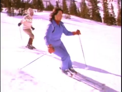The still features two individuals skiing on a snowy slope. One skier is wearing a blue outfit, actively navigating the slope with ski poles, while the other is further in the background, dressed in a lighter-colored outfit. The scene is set against a backdrop of snow-covered trees, emphasizing a winter sports theme related to physical fitness.
