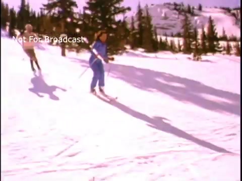 The still captures a vibrant winter scene where two individuals are skiing down a snowy slope, set against a backdrop of tall evergreens and distant mountains. The bright, clear sky adds to the cheerful atmosphere, emphasizing the theme of physical fitness and outdoor activity promoted in the PSA by John Denver. The individuals are dressed in colorful ski attire, reflecting the style of the era.
