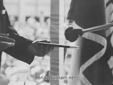 The still features a close-up of a hand holding a document, possibly a certificate or award, being presented at a podium. Behind it, a microphone is positioned with a flag visible in the frame. The setting suggests a formal event, likely a ceremony or announcement related to education, with a crowd in the background indicating public interest or participation.