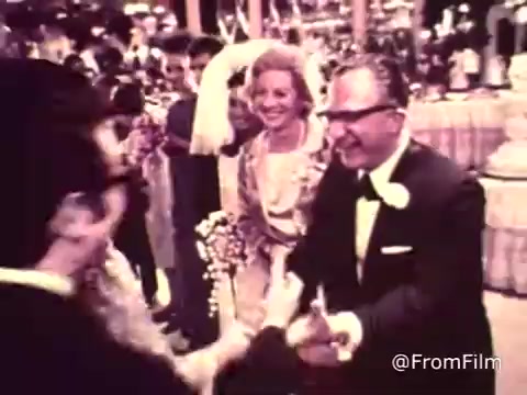 The 16mm still from the 'American Cancer Society PSA - 1969' depicts a lively wedding reception scene. People are dressed in formal attire, with a notable bride in a white wedding dress and veil. Guests are joyfully interacting, with smiling faces and gestures of celebration, like shaking hands or exchanging greetings. The background is adorned with decorations typical of festive occasions, creating a warm and inviting atmosphere. This segment likely serves to underscore themes of togetherness and joy, potentially contrasting with the PSA's message about cancer awareness.