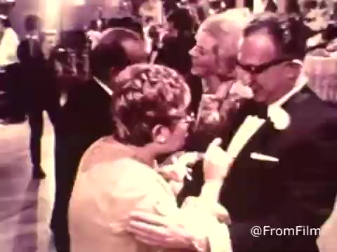 The 16mm still from the 1969 American Cancer Society PSA titled 'Wedding Reception' depicts a lively gathering, likely celebrating a wedding. The image features a group of people engaged in conversation and dancing, dressed in formal attire. A central focus is on a woman interacting warmly with another individual, while others appear to be enjoying the festivities in the background. The atmosphere is festive, capturing the essence of social gatherings of that era.