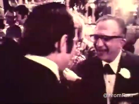 The still from the 16mm print of the 'American Cancer Society PSA - 1969' captures a lively wedding reception scene. You can observe a group of people engaged in conversation and celebration, dressed in formal attire. The atmosphere appears festive, with decorations characteristic of a wedding. The presence of a prominent figure, perhaps an elder, suggests a moment of applause or acknowledgment, indicative of the importance of community and support during that time. Overall, the still reflects a blend of joy and warmth typical of wedding gatherings.