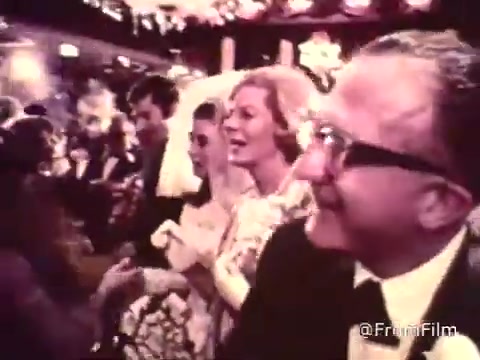 The still from the 1969 American Cancer Society PSA depicts a lively wedding reception scene. Guests are dressed in formal attire, with a bride in a white gown and veil prominently featured. The atmosphere is celebratory, with expressions of joy and engagement among the attendees. The image captures the essence of a social gathering, highlighting the importance of community and personal connections. This context ties into the PSA's messaging, focusing on awareness and support for cancer-related issues.