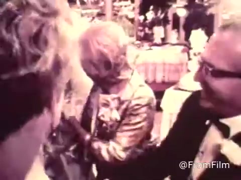 The still from the 16mm film titled 'American Cancer Society PSA - 1969' appears to capture a lively wedding reception scene. It features various guests engaging with each other, showcasing a blend of celebratory attire typical of that era. The image conveys a sense of joy and community, likely aiming to emphasize the importance of health awareness during a vibrant social moment. The colorful aesthetic and informal gathering highlight the personal aspect of the message from the American Cancer Society.