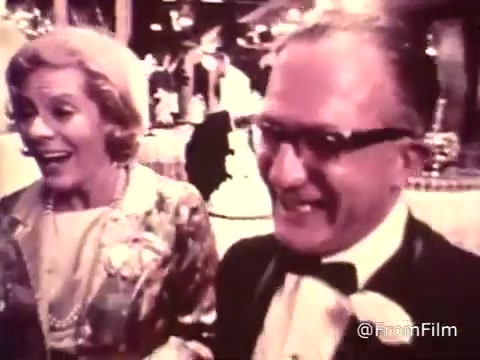 The still from the 'American Cancer Society PSA - 1969' depicts a cheerful wedding reception scene. In the image, a woman with styled hair and a patterned dress, adorned with pearls, appears to be laughing or enjoying a moment. Next to her, a man in a tuxedo and glasses is also smiling broadly, contributing to the festive atmosphere. The background features blurred elements typical of a reception setting, with decor and other attendees visible. The overall mood conveys joy and celebration, contrasting the serious message often associated with PSAs.