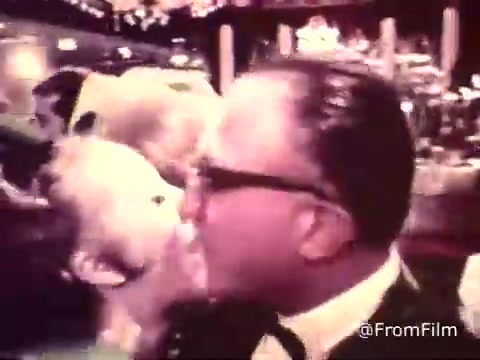 The still from the 16mm film 'American Cancer Society PSA - 1969' depicts a wedding reception scene. In the foreground, an older gentleman wearing glasses is engaged with a young child who is playfully interacting with him, likely giving him a kiss. The setting appears vibrant and festive, consistent with a celebratory atmosphere typical of wedding receptions. Background details suggest a lively environment, with blurred figures and decorations enhancing the joyful mood of the occasion.