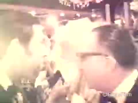 The still from the 1969 American Cancer Society PSA features a wedding reception scene captured on 16mm film. The image portrays two men engaged in conversation, with a soft glow and slightly blurred focus typical of older film formats. The background suggests a lively celebratory atmosphere, consistent with the joyful context of a wedding. The overall tone combines both social interaction and a sense of warmth, reflective of the era's aesthetic.