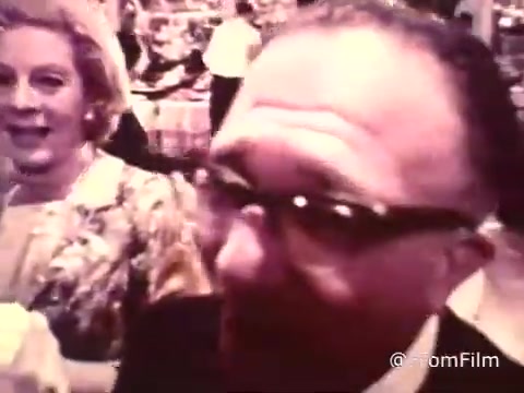 The still from the American Cancer Society PSA from 1969 features a lively wedding reception scene. It showcases two individuals: a man wearing glasses smiling directly at the camera and a woman beside him with a joyful expression. The background reveals festive decorations and food typical of such gatherings. The warm colors capture the celebratory atmosphere, highlighting a moment of social interaction during the event.