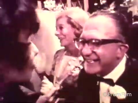 The still from the 1969 American Cancer Society PSA features a lively wedding reception scene. In the foreground, a man wearing glasses and a tuxedo is smiling while engaged in a conversation with a woman. Behind them, another woman in a formal outfit appears to be enjoying the moment. The setting is decorated and festive, typical of a wedding celebration, subtly blending the themes of community and support that are central to the PSA’s message. The image likely aims to highlight awareness and solidarity in the fight against cancer.