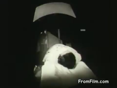 The still from the 16mm PSA 'Cobalt Bomb' depicts a patient undergoing cancer treatment in a hospital setting from 1954. The image shows the patient lying on a treatment table, possibly receiving radiation therapy. The lighting is dim, emphasizing the seriousness of the medical procedure, which reflects the era’s advancements in cancer treatment and public health messaging.