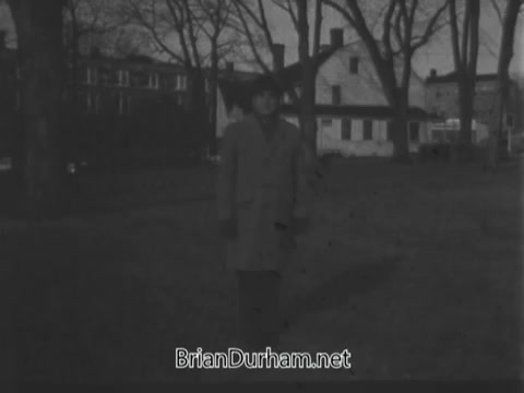 The still from the 1968 'Guideposts Magazine' commercial features a person standing in a park-like setting, surrounded by bare trees. The background includes a few buildings, and the overall mood seems somber, likely due to the black-and-white film. The individual is dressed in a coat, suggesting a cool climate. The focus appears to be on the person, emphasizing a moment of introspection or connection.