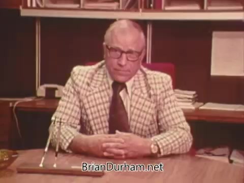 The 16mm still features a formal-looking man seated at a desk, dressed in a plaid suit. He appears to be addressing an audience, likely conveying a message related to the political advert against Issue Six in Ohio, which pertains to nuclear power. The background includes shelves filled with papers, suggesting a professional setting. His serious demeanor indicates the importance of the topic at hand.