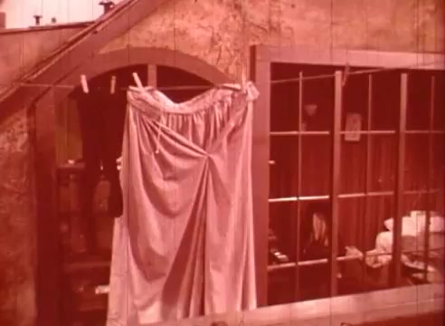 The still from 'Erik Satie and the King of the Beans - Part II' features a rustic scene with a large garment hanging on a line outside a window. The background shows a stone wall and several windows with visible curtains, creating an intimate, domestic atmosphere. The color tone is warm, possibly sepia, enhancing a nostalgic or whimsical feel. The image suggests simplicity and everyday life, aligning with the artistic and surreal themes of the film.