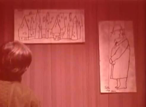 The 16mm still from 'Erik Satie and the King of the Beans - Part II' features a character facing a wall adorned with two drawings. One illustration depicts whimsical, abstract houses with varying shapes and sizes, while the other shows a cartoonish figure in a coat and hat. The scene has a reddish tint, contributing to a surreal atmosphere, reflecting the whimsical nature of the narrative.