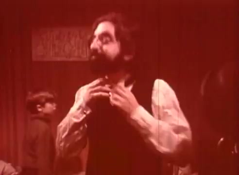 The still from 'Erik Satie and the King of the Beans - Part II' features a man with a beard adjusting his collar, dressed in period attire. He appears thoughtful and is set against a backdrop that suggests a cozy, perhaps whimsical interior. A boy in the background adds to the scene, enhancing the playful tone of the narrative. The warm red tint gives a unique, nostalgic feel to the image.
