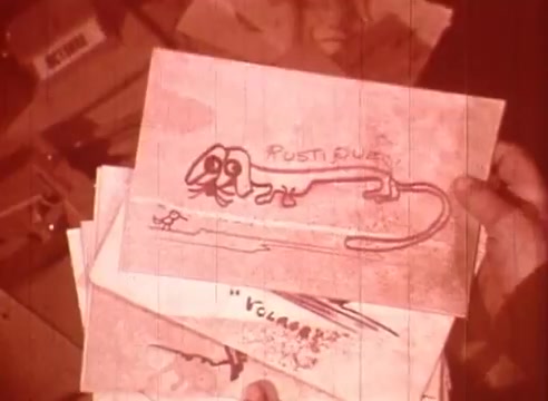 The still features a hand holding a stack of illustrated drawings, prominently displaying a cartoonish dog with exaggerated features. The drawings seem playful and whimsical, suggesting a lighthearted tone. Text elements like 'POSTI DUE' and “VOLARE” are visible, hinting at a narrative or thematic connection related to music or comedy. The overall visual style has a warm, faded quality typical of vintage film, contributing to its nostalgic atmosphere.