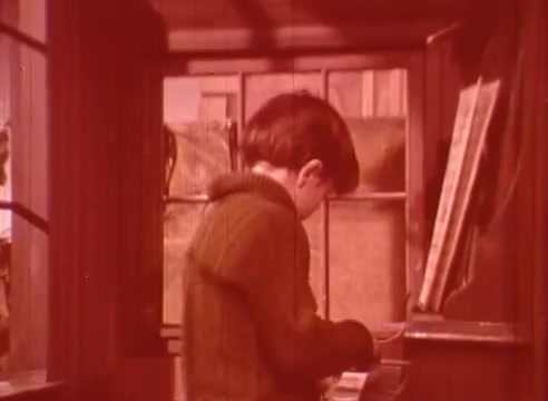 The still depicts a young boy playing a piano in a cozy, sunlit interior, surrounded by windows. He is focused on the keys, with his back turned to the viewer. The warm red tint adds a nostalgic and whimsical atmosphere, enhancing the enchanting mood of the scene from 'Erik Satie and the King of the Beans - Part II.'