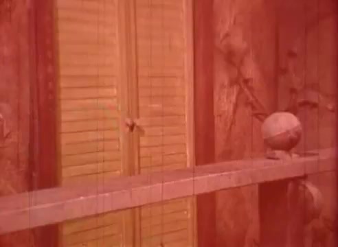 The still features a warm, reddish tint and presents a close-up view of a wooden ledge with a round object on it, possibly a ball. In the background, there are shutters, adding a sense of depth. The scene evokes a whimsical, surreal atmosphere characteristic of Erik Satie's artistic style.