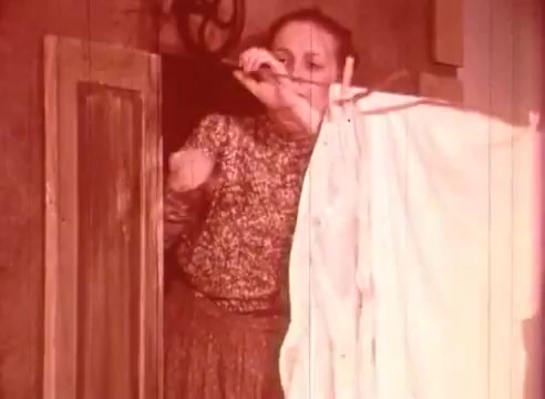 The still from 'Erik Satie and the King of the Beans - Part II' depicts a woman in a patterned blouse and skirt, standing at a doorway. She appears to be holding a white cloth, possibly a curtain or garment, and is engaged in a thoughtful or playful manner. The image has a warm, reddish tint, evoking a nostalgic feel typical of 16mm film. The setting suggests an intimate or whimsical atmosphere, aligning with the artistic themes often associated with Erik Satie's work.