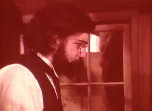 The 16mm still from 'Erik Satie and the King of the Beans - Part II' features a man with a beard and glasses, dressed in period attire, staring pensively towards a window or door. The image has a warm, reddish tint, enhancing its nostalgic atmosphere. The scene suggests introspection or contemplation, fitting the imaginative theme of the film centered around the composer Erik Satie.