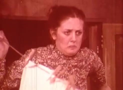 The still from 'Erik Satie and the King of the Beans - Part II' features a woman with a distressed expression, holding a garment and a rod, suggesting a moment of confusion or alarm. The warm color palette enhances the emotional intensity of the scene, hinting at a comedic or surreal narrative.