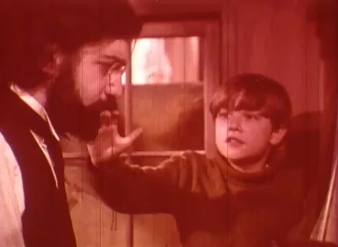 The still from 'Erik Satie and the King of the Beans - Part II' features two characters engaged in an intense conversation. One character, a man with a beard and glasses, appears serious, while the boy, dressed in a sweater, is animated and gesturing with his hand. The warm, reddish tint of the image creates a nostalgic or whimsical mood, enhancing the emotional atmosphere of the scene.