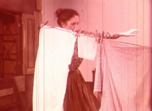 The still from 'Erik Satie and the King of the Beans - Part II' depicts a woman hanging laundry. She is wearing a long skirt and appears engaged in her task, with a focus on the movement of her arms as she hangs pieces of clothing. The warm, muted tones of the image suggest a nostalgic or whimsical atmosphere, consistent with the creative style of the film. The setting features simple interior elements, adding to the everyday nature of the scene.