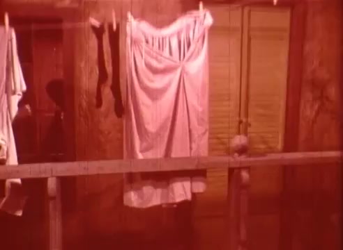 The still from 'Erik Satie and the King of the Beans - Part II' features a rustic interior scene, with clothes hanging on a line. The colors have a warm, reddish tint, creating a nostalgic or dreamlike atmosphere. The background suggests an old wooden structure, enhancing the intimate and domestic feel of the moment. The simplicity of the scene reflects a focus on everyday life, resonating with Satie's themes of subtlety and the ordinary.