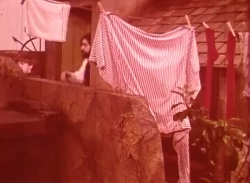 The still from 'Erik Satie and the King of the Beans - Part II' illustrates a whimsical scene featuring a character near a rustic setting. A striped garment hangs prominently on a line, adding a playful contrast to the earthy tones of the background. The character's facial expression hints at curiosity or contemplation, contributing to the overall surreal atmosphere of the film. The setting feels intimate and slightly nostalgic, evoking a sense of simplicity amidst the quirky narrative.