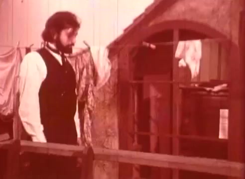 The still from 'Erik Satie and the King of the Beans - Part II' features a man dressed in period attire, including a vest and shirt. He stands in front of a rustic structure, which resembles a small building or house. The setting is enhanced by hanging garments, adding to the atmosphere. The color tone is warm, giving the scene a vintage feel.