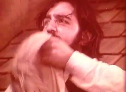 The still from 'Erik Satie and the King of the Beans - Part II' features a man with long hair and glasses, expressing intense emotion as he interacts with a cloth or piece of fabric. The warm color palette adds a vintage feel, reflecting the film's stylistic choices. The setting appears rustic, enhancing the whimsical atmosphere typical of works inspired by Satie's music.